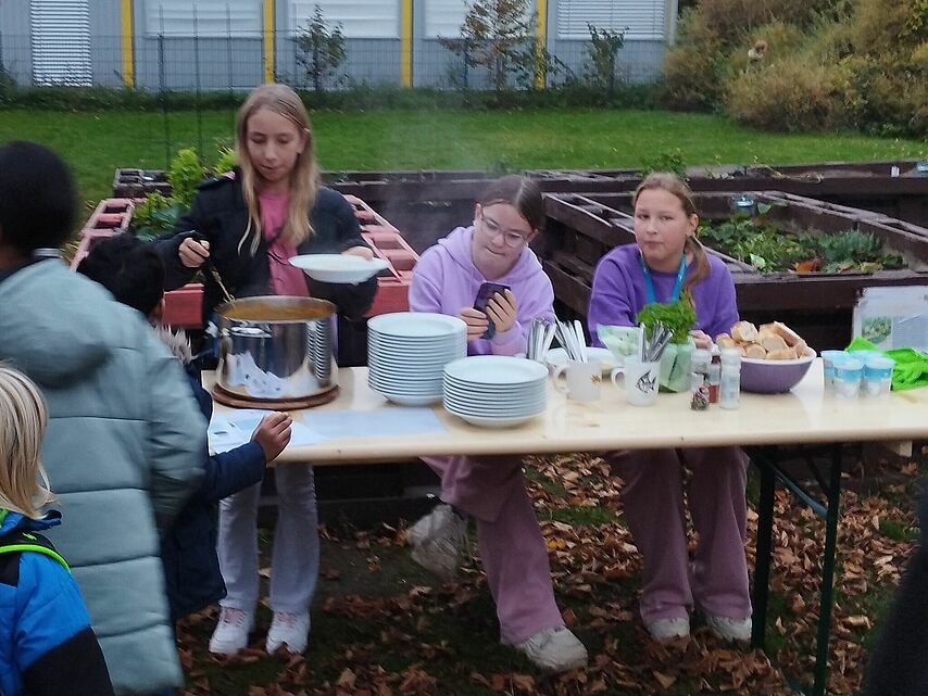 Auf dem Bild ist eine kleine Outdoor-Veranstaltung zu sehen. Drei Mädchen stehen an einem Tisch, der mit Tellern, Bechern und anderen Utensilien gedeckt ist. Eine der Mädchen serviert eine warme Mahlzeit aus einem großen Topf. Einige Kinder, die in winterlicher Kleidung wie Jacken und Mützen gekleidet sind, stehen in der Nähe des Tisches und warten darauf, ihr Essen zu bekommen. Der Hintergrund zeigt einen Gartenbereich mit Hochbeeten und bunten Herbstblättern, die den Boden bedecken