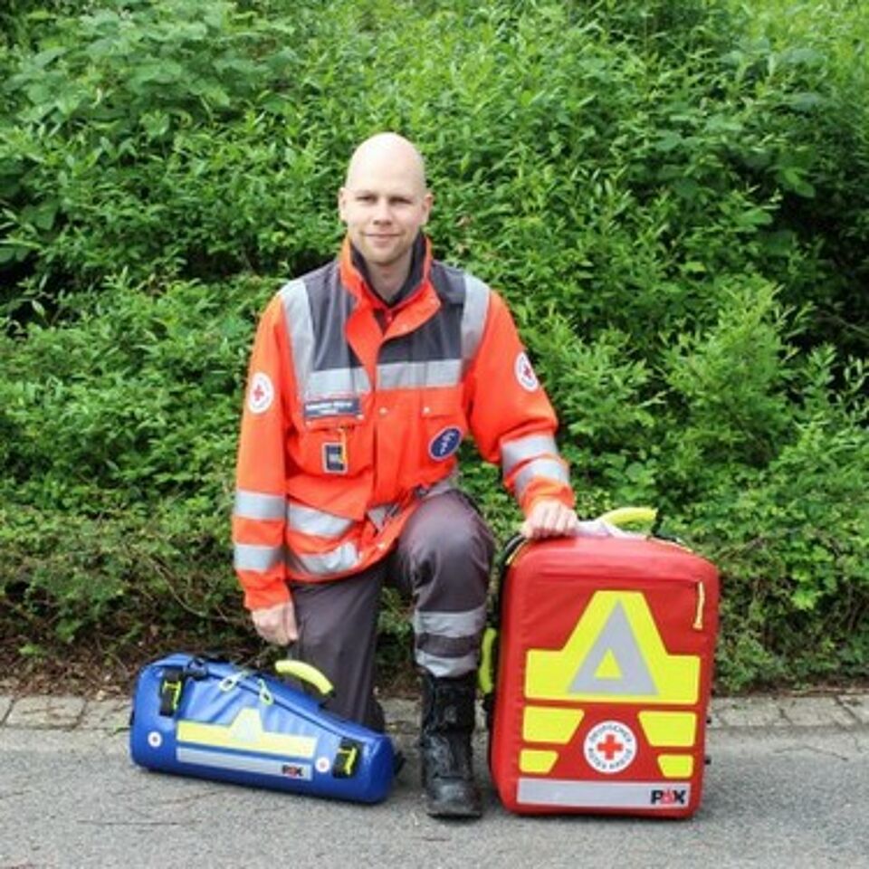 Eine Person in Einsatzkleidung kniet neben einem Notfallrucksack. Im Hintergrund befindet sich grüne Vegetation.