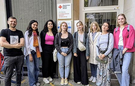 Gruppenbild von acht Personen vor einem Hauseingang, an dem ein Schild vom DRK mit den Angeboten "Freiwilligendienste, Migration und Sozialberatung" hängt