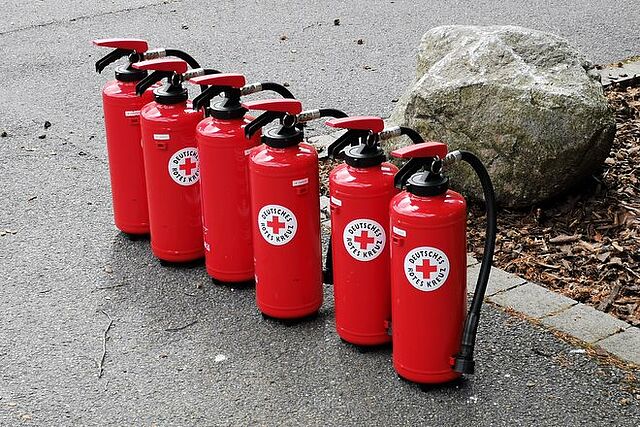 Sechs rote Feuerlöscher mit einem DRK-Logo stehen in einer Reihe auf dem Boden.