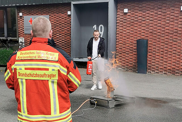 EIne Person löscht mit einem Feuerlöscher ein Feuer, vorne im Bild steht eine Person mit dem Rücken zur Kamera und trägt eine Einsatzjacke mit der Beschriftung "Brandschutzbeauftragter"