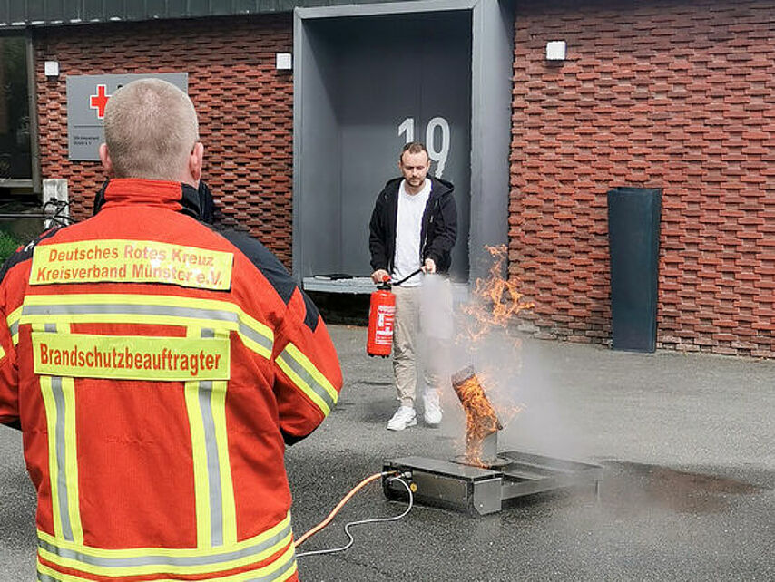 EIne Person löscht mit einem Feuerlöscher ein Feuer, vorne im Bild steht eine Person mit dem Rücken zur Kamera und trägt eine Einsatzjacke mit der Beschriftung "Brandschutzbeauftragter"