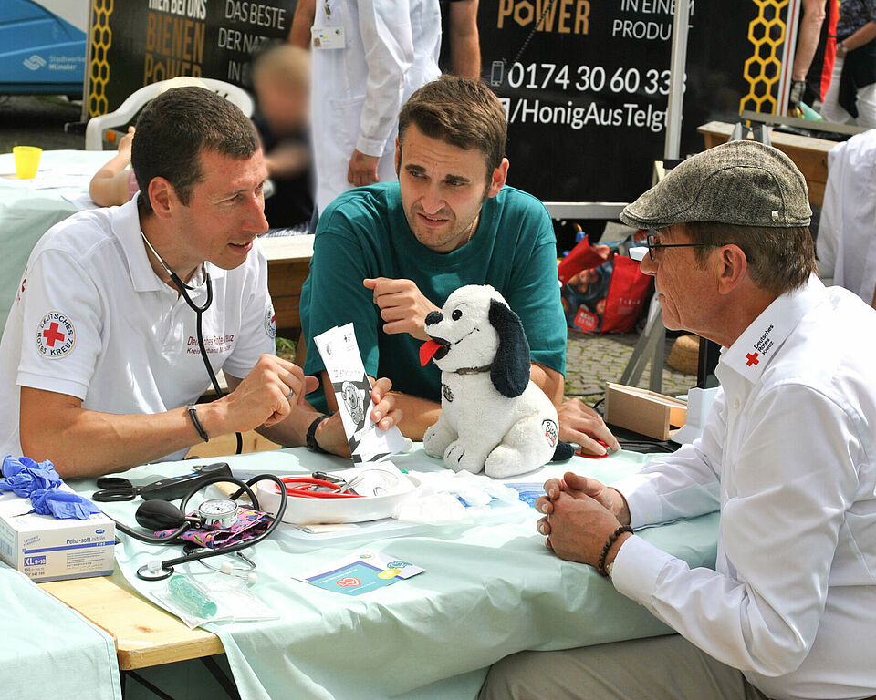 Drei Personen in JRK- und DRK-Kleidung sitzen an einem Tisch mit Utensilien der Teddyklinik