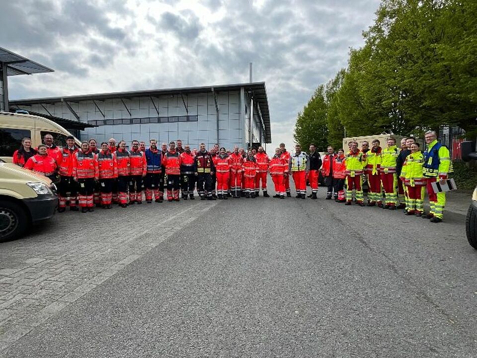 Betreuungsdienst Gruppenbild Gruppenbild von Personen in Einsatzkleidung, im Hintergrund ist eine Messehalle zu sehen, an den Seiten stehen jeweils Fahrzeuge