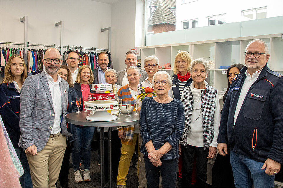 10 Jahre Kleiderladen Gruppenbild im Kleiderladen, in der Mitte steht ein Stehstisch mit Torte und Sekt, im Hintergrund Regale des Ladens