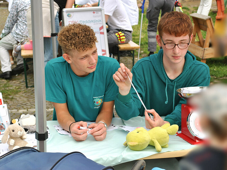 Zwei Jugendliche in JRK-Shirts sitzen an einem Tisch mit Kuscheltieren und Utensilien der Teddyklinik