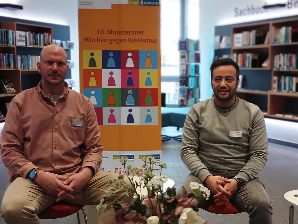 Zwei Personen sitzen vor dem Aufsteller der Wochen gegen Rassismus, im Hintergrund Bücherregale