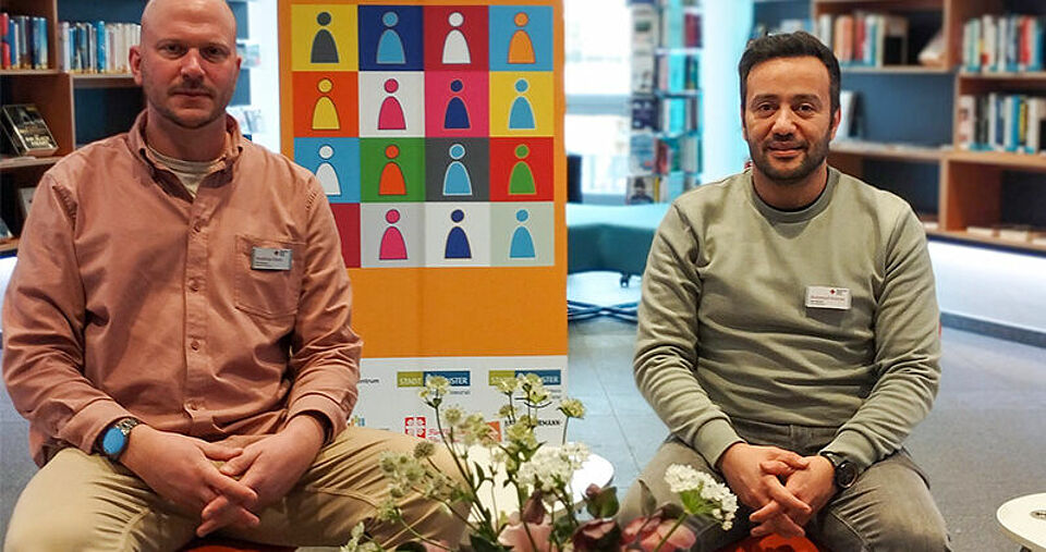 Zwei Personen sitzen vor dem Aufsteller der Wochen gegen Rassismus, im Hintergrund Bücherregale