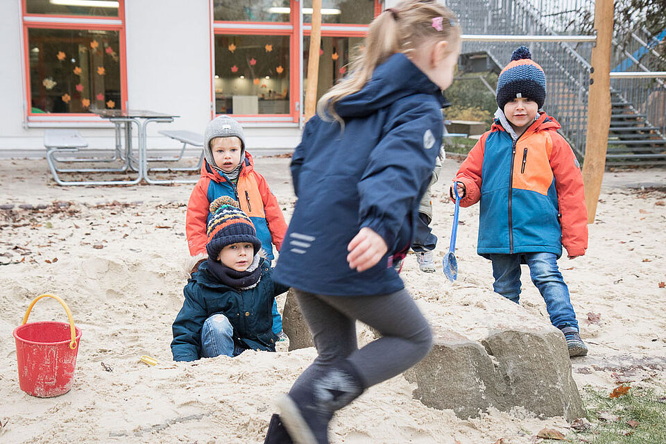 Vier Kinder in Winterkleidung spielen in einem Sandkasten, im Hintergrund ist ein Gebäude zu sehen