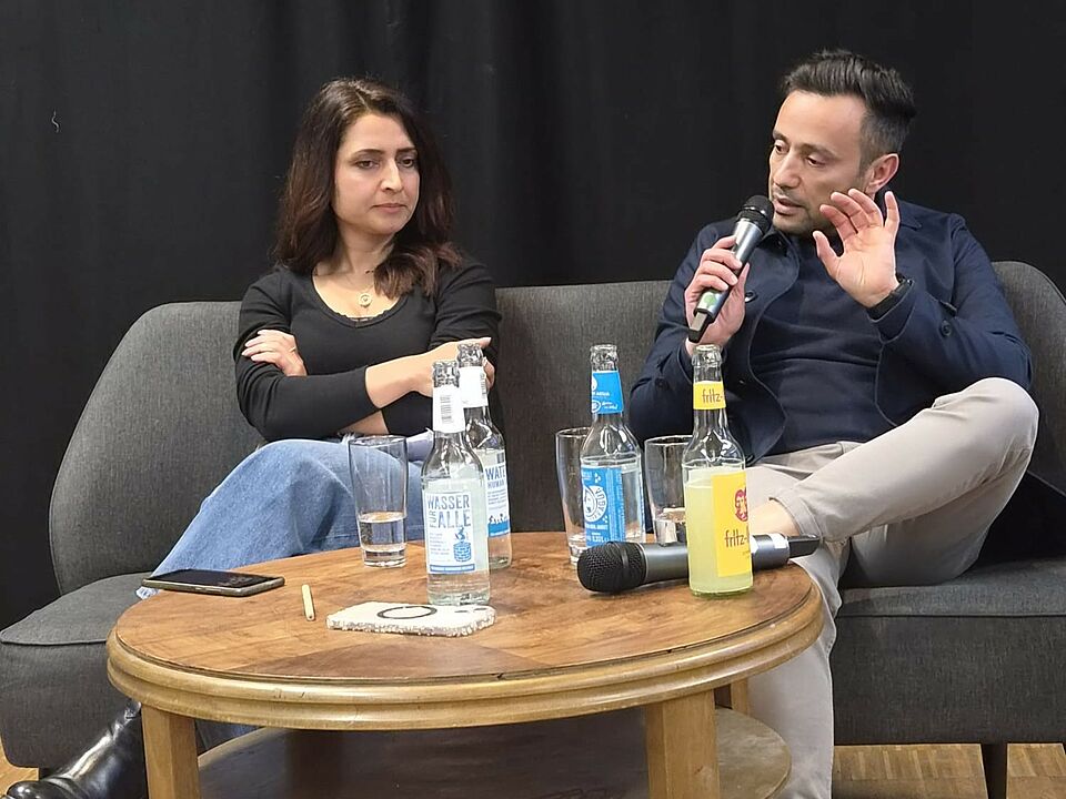 Eine Frau und ein Mann sitzen auf einem Sofa, der Mann hat ein Mikrofon in der Hand. Vor ihnen steht ein Holztisch mit Getränken.