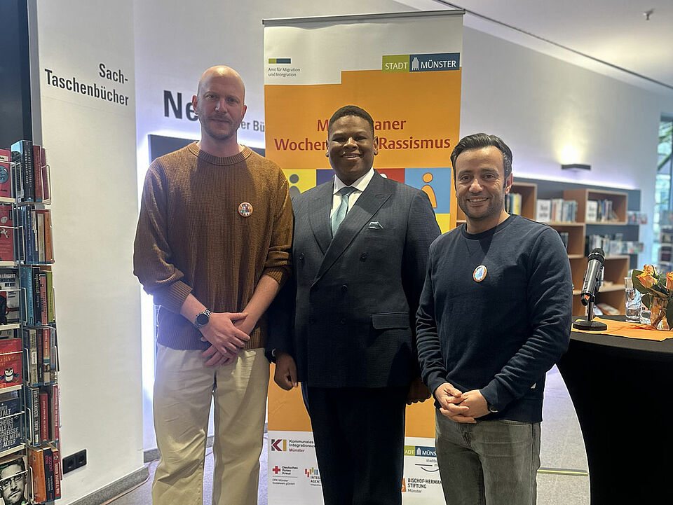 Drei Männer vor einem Banner der Wochen gegen Rassismus, im Hintergrund Bücherregale