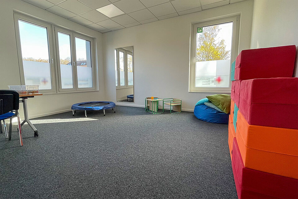 Ein Raum mit Fenstern, bunten Schaumstoffwürfeln, einem Trampolin und einem Tisch mit Stühlen.