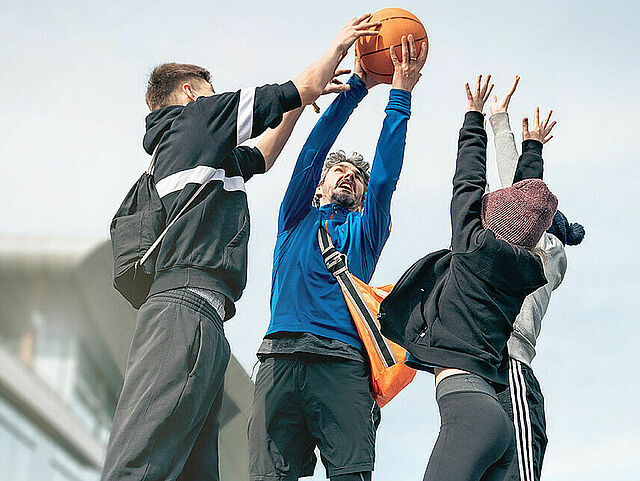 Drei Personen strecken sich nach einem Basketball in der Luft