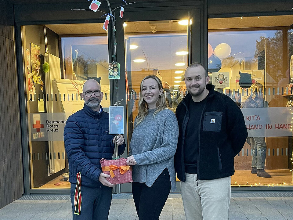 Apfelbaum Drei Personen stehen vor einem Gebäude mit einer Glasfront und DRK-Logo. Sie halten einen kleinen Baum im Topf.