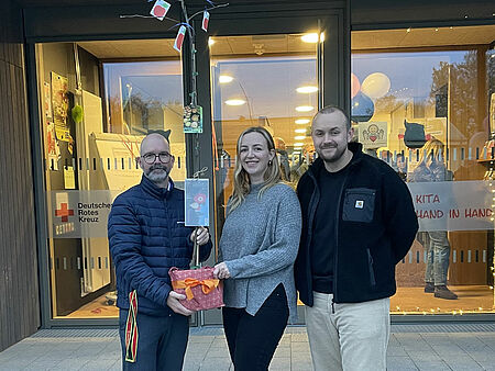 Drei Personen stehen vor einem Gebäude mit einer Glasfront und DRK-Logo. Sie halten einen kleinen Baum im Topf.