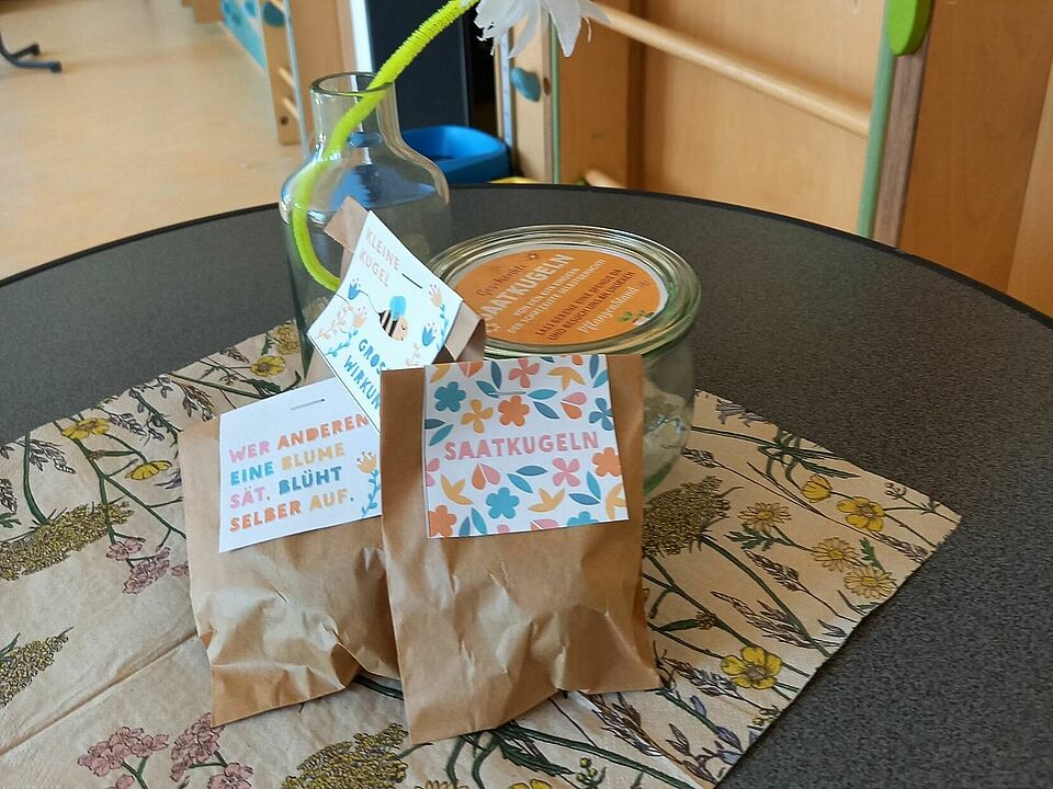 Ein Tisch auf dem neben einer Blume kleine Packtüten stehen, auf denen auf einem bunten Schild "Saatkugeln" steht.