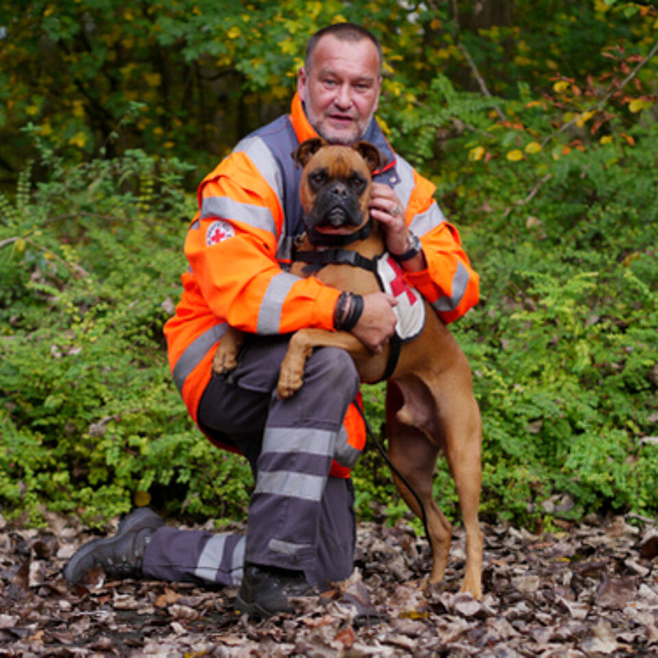 Eine Person in Einsatzkleidung kniet neben einem Hund. Im Hintergrund befindet sich grüne Vegetation.