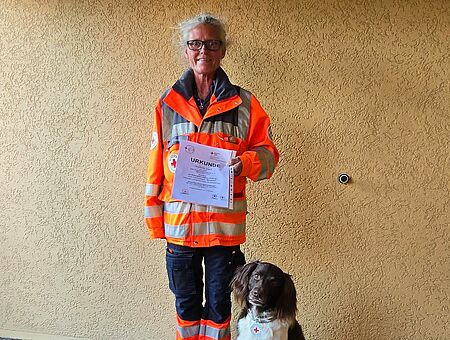 Eine Person in Einsatzkleidung steht vor einer hellen Wand. In der rechten Hand hält sie eine Urkunde. Zu ihrer linken Seite sitzt ein mittelgroßer braun-weißer Hund mit einem Halsband, an dem eine kleine DRK-Plakette befestigt ist.