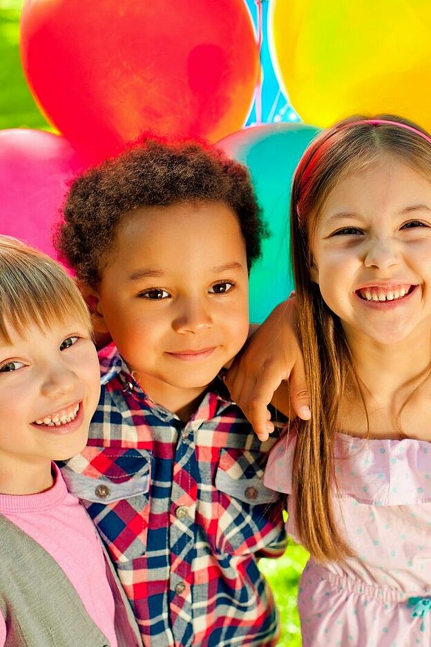 Vier Kinder stehen eng beieinander und haben die Arme umeinander gelegt. Im Hintergrund sind viele bunte Luftballons zu sehen