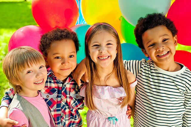 Vier Kinder stehen eng beieinander und haben die Arme umeinander gelegt. Im Hintergrund sind viele bunte Luftballons zu sehen