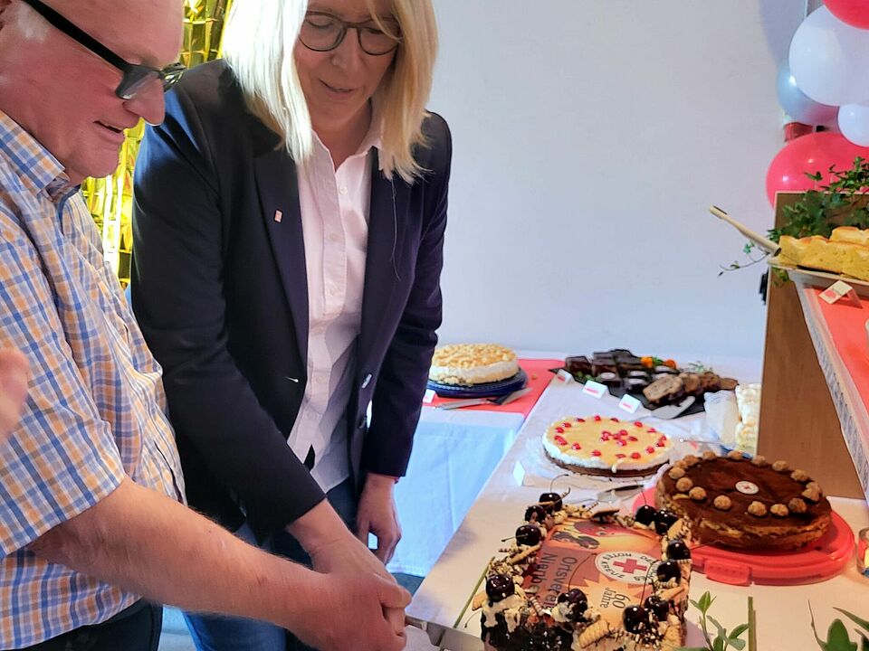 Eine Frau und ein Mann schneiden gemeinsam eine Torte an. Im Hintergrund sind weitere Torten und weiß-rote Luftballons zu sehen.