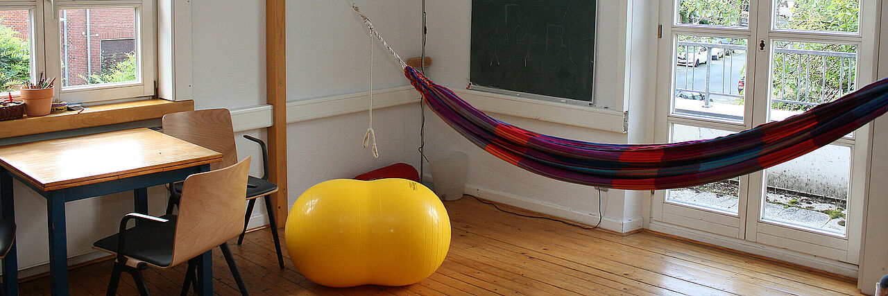 Kinderschutzambulanz Ein Raum mit Holzfußboden, einem Tisch mit zwei Stühlen, einem großen gelben Gymnastikball und einer bunten Hängematte. An der Wand hängt eine Tafel, und durch die Fenster fällt Tageslicht in den Raum.