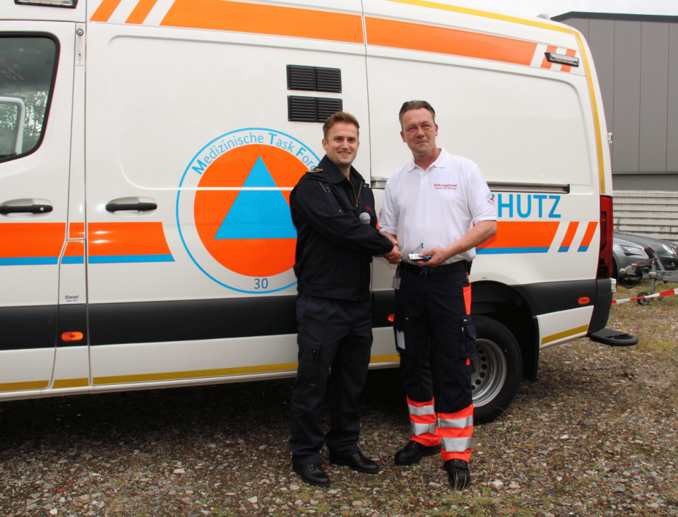 Zwei Männer stehen vor einem Einsatzfahrzeug. Person in Feuerwehreinsatzkleidung überreicht Person in Rotkreuzeinkleidung einen Schlüssel.