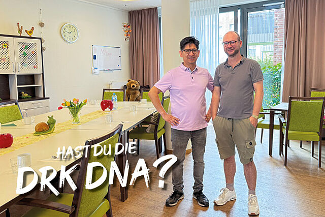 Zwei Männer stehen neben einem großen Esstisch mit grünen Stühlen im Gemeinschaftsbereich einer Wohngemeinschaft.