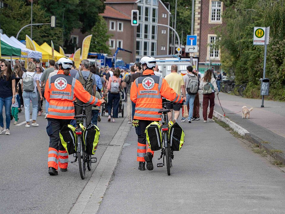 Zwei Personen in Einsatzkleidung sind von hinten zu sehen und schieben zwei Fahrräder. Im Hintergrund ist die Blaulichmeile mit vielen Pavillions und Menschen zu sehen