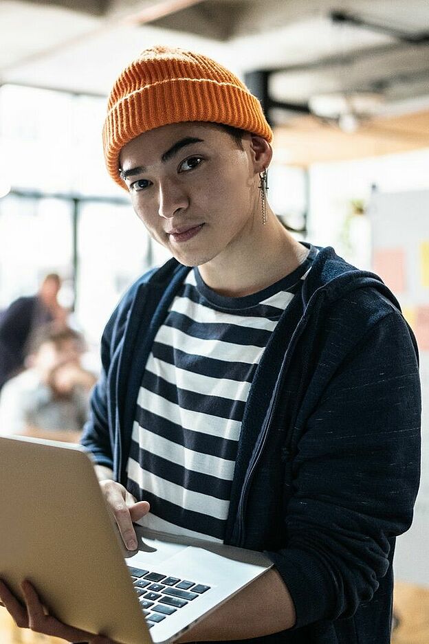 Eine Person mit orangefarbener Mütze und gestreiftem Oberteil hält ein aufgeklapptes Laptop in den Händen. Im Hintergrund sitzen mehrere weitere Personen an Tischen und beschäftigen sich mit verschiedenen Aktivitäten. An der Wand steht ein Whiteboard mit unleserlicher Beschriftung.