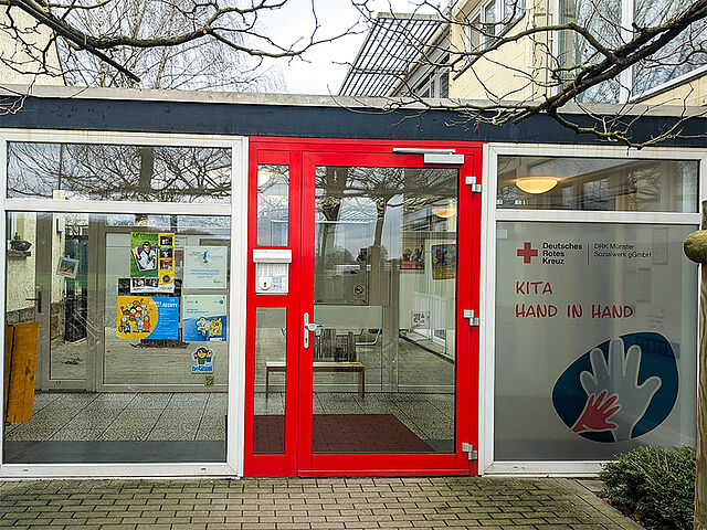 Eingang eines Gebäudes mit roter Tür und großen Glasfenstern. Die Tür verfügt über einen Briefschlitz und eine Gegensprechanlage. Rechts neben der Tür befindet sich ein Schild mit dem Logo des Deutschen Roten Kreuzes und der Aufschrift ‚KITA HAND IN HAND‘. Durch die Fenster sind Poster an den Wänden und Möbel im Innenraum zu erkennen.
