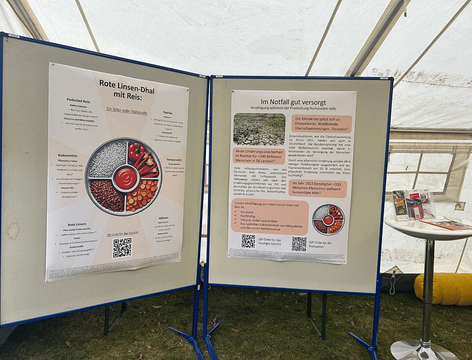 Zwei Stellwände in einem Zelt, wo jeweils ein Plakat dran hängt. Links ist ein Rezept für Linsendaal und rechts zu Ernährung in Notfallsituationen