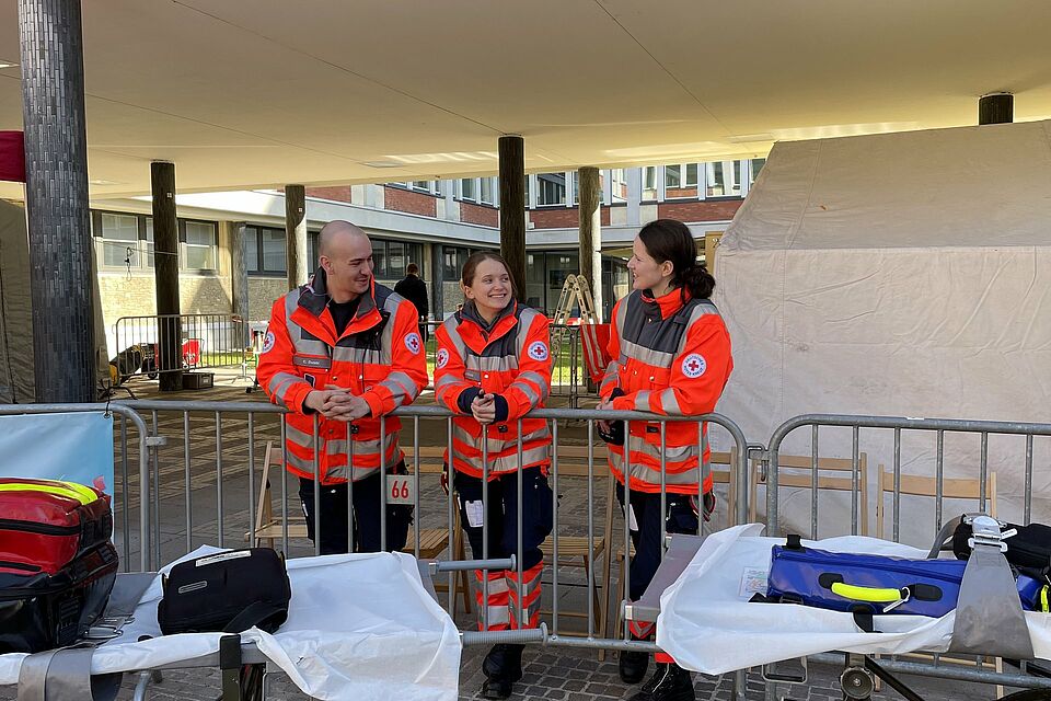 Drei Personen in Einsatzkleidung stehen an einem Absperrgitter. Davor stehen mobile Tragen und Erste Hilfe Equipment. Im Hintergrund befinden sich Zelte.