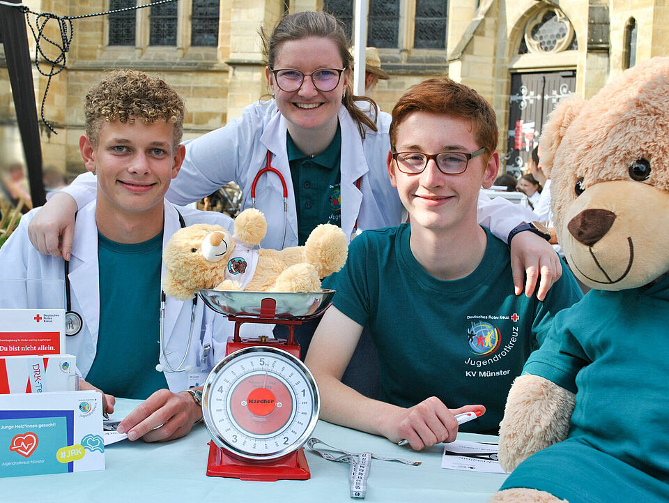 Drei Personen sitzen und stehen Arm in Arm hinter einem Tisch. Auf dem Tisch befinden sich Teddys und eine Waage und Infomaterialien des Rotes Kreuzes