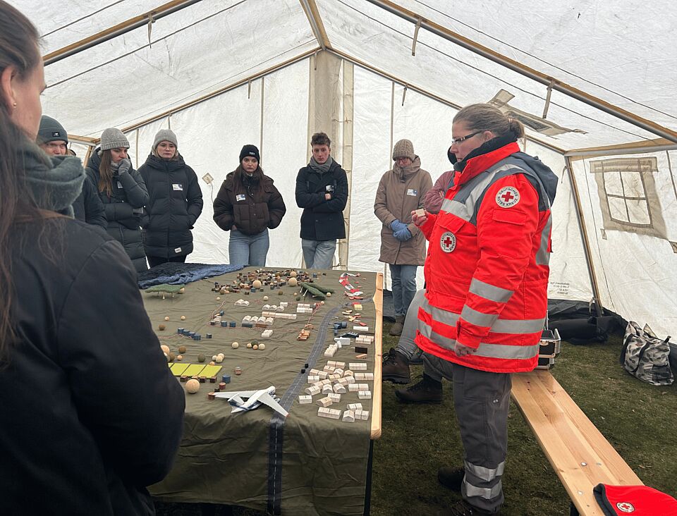 Eine Gruppe steht in einem Zelt, eine Person trägt Einsatzkleidung. In der Mitte befindet sich ein Tisch mit vielen Miniaturzelten.