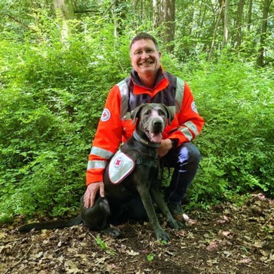 Eine Person in Einsatzkleidung kniet neben einem Hund. Im Hintergrund befindet sich grüne Vegetation.
