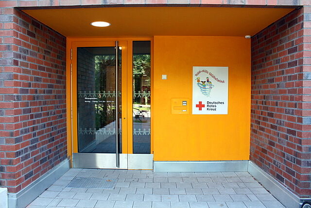 Eingang eines Gebäudes mit einer orangefarbenen Wand und einer Glastür. An der Wand befindet sich ein Schild mit dem Schriftzug "Deutsches Rotes Kreuz" und einem roten Kreuz-Symbol. Daneben ist ein Briefkasten angebracht. Die angrenzenden Wände bestehen aus roten Ziegelsteinen.
