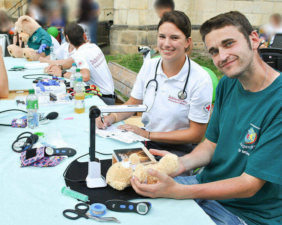 Mehrere Personen sitzen an einem langen Tisch auf dem Utensilien der Teddyklinik liegen. Im Fokus stehen zwei Personen, die vor dem Gerät, dass das röntgen simuliert, sitzen und in die Kamera schauen.