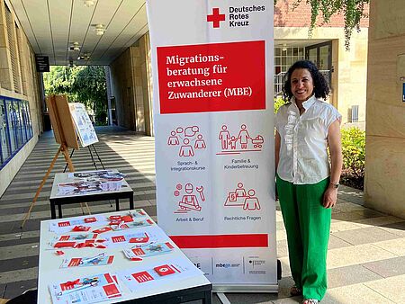 Eine Frau steht neben einem Tisch mit Infomaterial und einem Roll-Up zur Migrationsberatung.