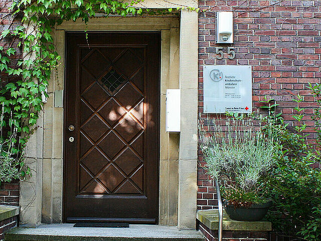 Eingangsbereich eines Backsteingebäudes mit brauner Tür und Treppe, flankiert von zwei Pflanzkübeln mit hohen Pflanzen. Über der Tür wächst Efeu an der Wand. Links befindet sich ein Fenster mit weißen, teils geöffneten Fensterläden. Rechts blühen pinke Blumen. Ein Schild mit der Hausnummer „55“ hängt neben der Tür.