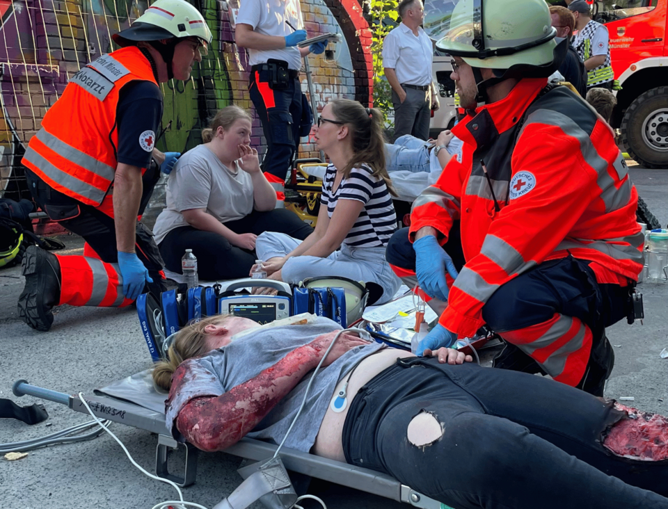 Im Vordergrund liegt eine verletzte Person mit blutenden Wunden auf einer Trage, neben ihr kniet eine Einsatzkraft, im Hintergrund sitzen mehrere Personen auf dem Boden und eine Einsatzkraft spricht mit diesen Personen.
