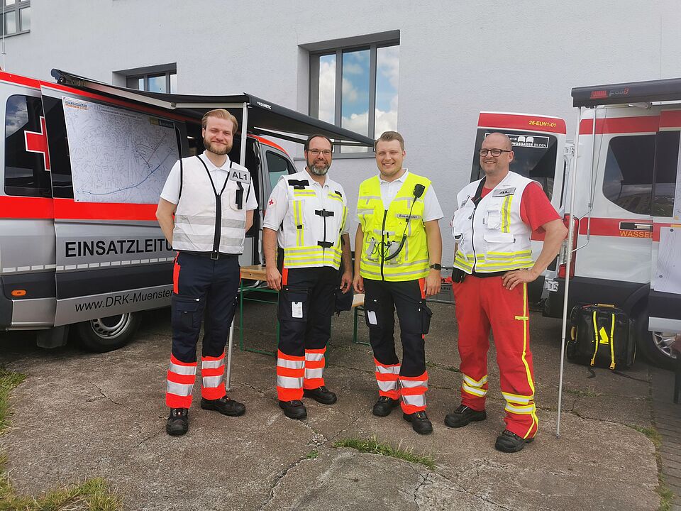 Vier Personen mit Einsatzkleidung und Westen stehen zwischen den Einsatzleitungswägen von DRK und DLRG