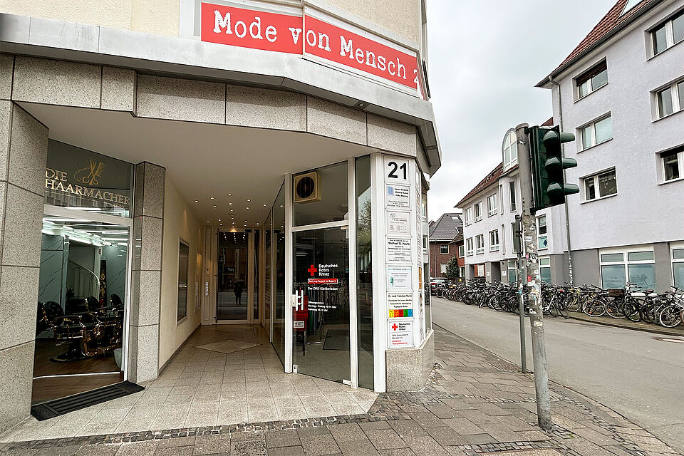 Straßenecke mit einem weißen Hauseingang mit DRK-Logo und Schriftzug. Mittig geht ein Gang zu einer weiteren Tür. Link ist der Eingang eines Friseurladens