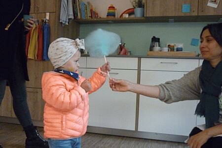 Eine Person gibt einem Kleinkind eine Zuckerwatte am Stiehl, im Hintergrund ist eine Küche zu sehen
