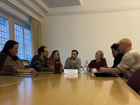 Sieben Personen sitzen in einem Halbkreis um einen Tisch herum