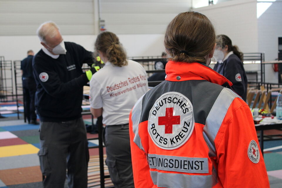 Mehrere Personen tragen Uniformen des Deutschen Roten Kreuzes. Im Vordergrund ist eine Person mit einer leuchtend orangefarbenen Jacke zu sehen, auf der „DEUTSCHES ROTES KREUZ“ und „RETTUNGSDIENST“ steht. Im Hintergrund trägt eine andere Person ein weißes Shirt mit der Aufschrift „Deutsches Rotes Kreuz Kreisverband Münster“. Die Szene spielt sich in einem Innenraum ab, möglicherweise in einer großen Halle mit buntem Bodenbelag und es werden Stockbetten aufgebaut.