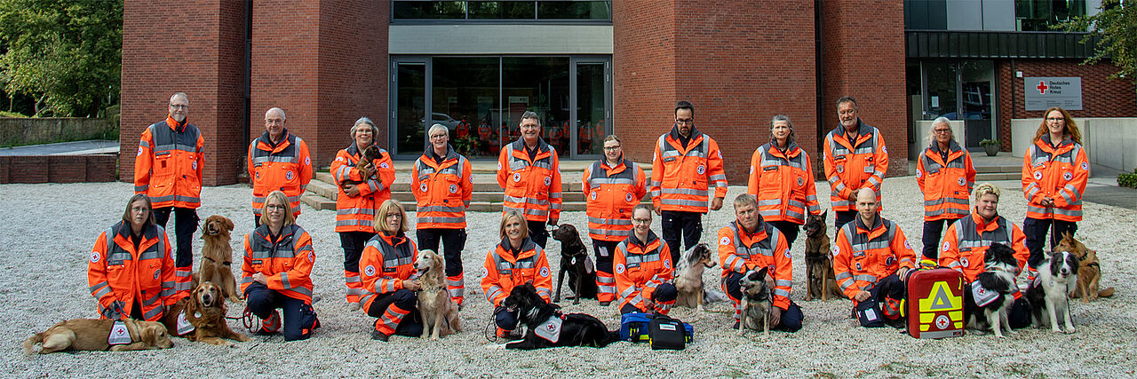 Eine Gruppe von Personen in Einsatzkleidung steht vor einem Backsteingebäude. Sie werden von mehreren Hunden begleitet, die teils sitzen oder liegen. Auf dem Boden liegen medizinische Ausrüstungsrucksäcke. Im Hintergrund sind große Glasfenster und -türen zu sehen, rechts befindet sich ein Schild mit dem Rotkreuz-Symbol.