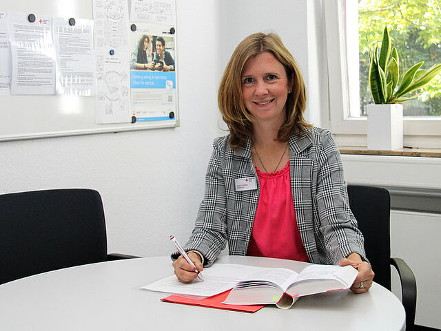Person sitzt an einem Tisch mit aufgeschlagenem Buch und Unterlagen und schreibt mit einem Stift. Im Hintergrund eine Pinnwand mit Aushängen sowie ein Fenster mit Blick ins Grüne