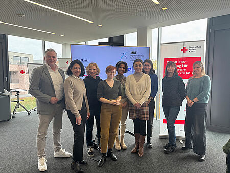 Eine Gruppe von neun Personen steht vor einem Banner und einem großen Bildschirm in einem Raum mit Glasfronten.