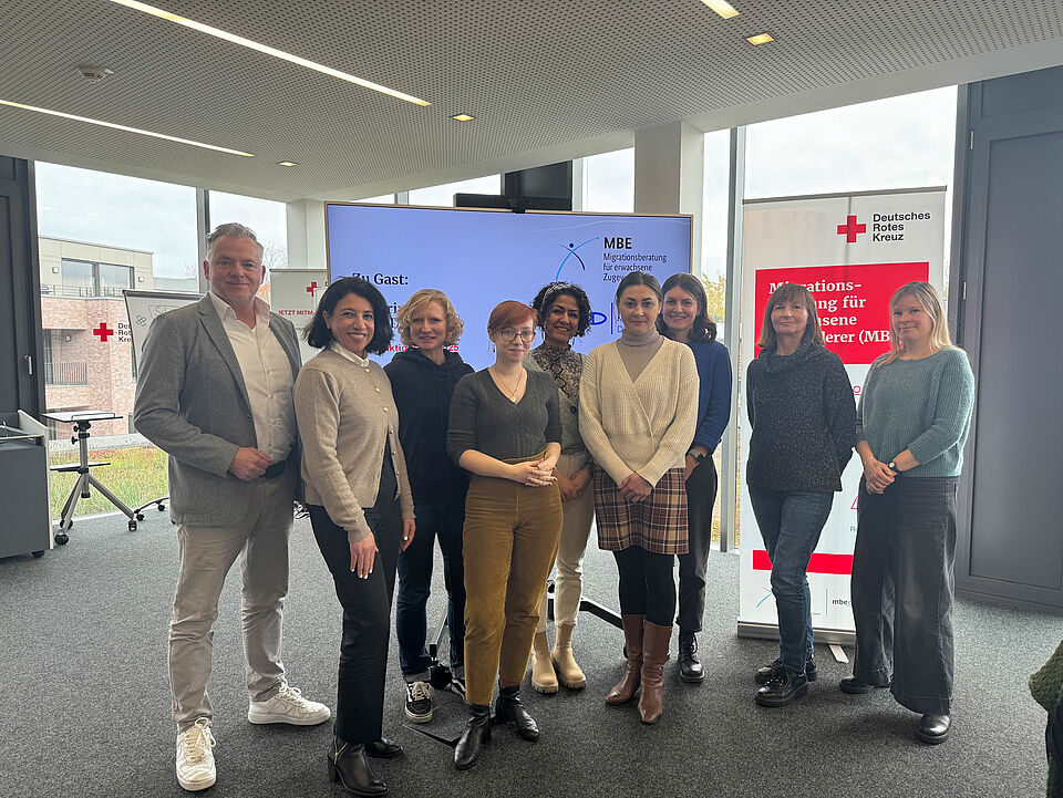 Gruppenbild Eine Gruppe von neun Personen steht vor einem Banner und einem großen Bildschirm in einem Raum mit Glasfronten.